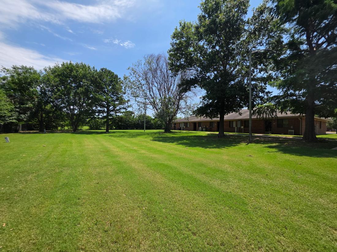 A wide, freshly mowed lawn shaded by large trees behind a row of single-story brick apartments